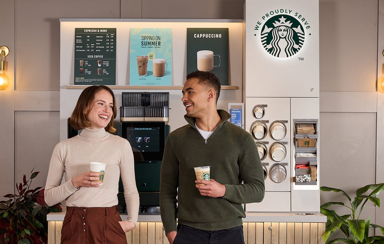 Twee mensen staan te praten terwijl ze We Proudly Serve Starbucks koffiebekers vasthouden, voor een zelfbedieningsstation
