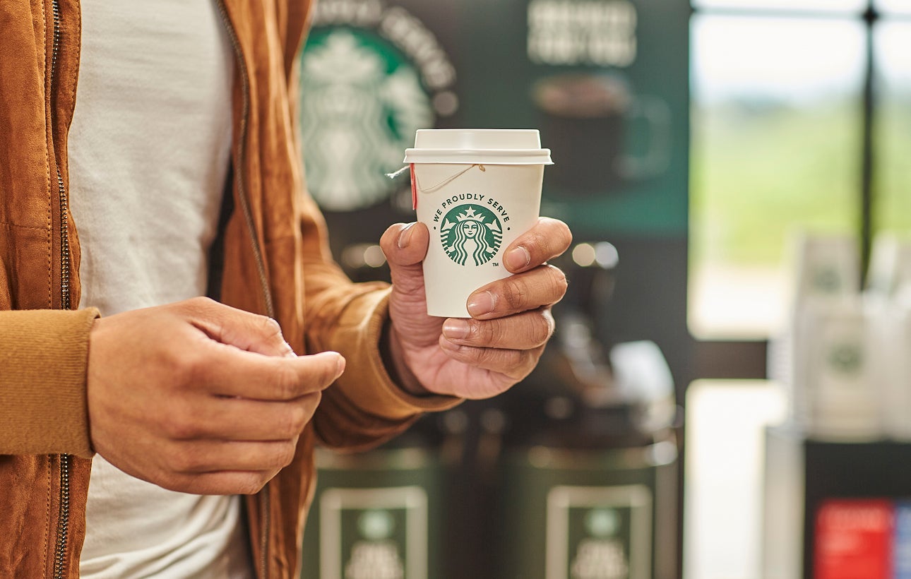 Een persoon die een kop thee vasthoudt in een We Proudly Serve Starbucks take-away beker
