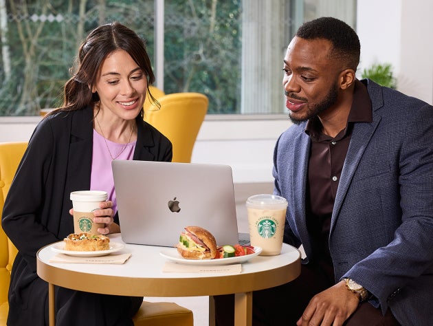 Twee mensen zitten voor een laptop met We Proudly Serve Starbucks koffie en broodjes op tafel