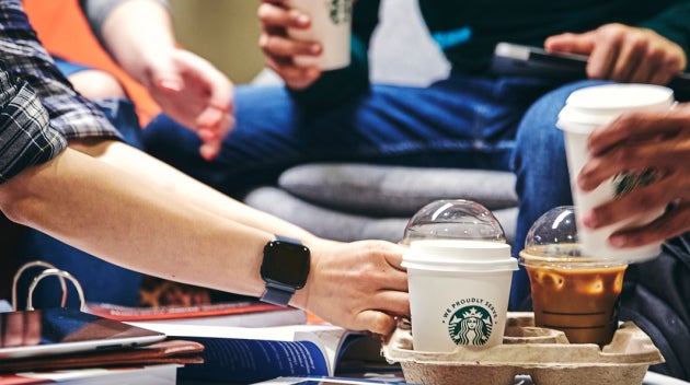 Een groep studenten zit rond een tafel met boeken en schriften; een van hen pakt een We Proudly Serve Starbucks koffie van de
