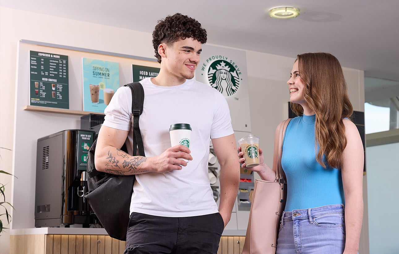 Un homme et une femme debout devant une machine à café autonome en libre-service We Proudly Serve Starbucks