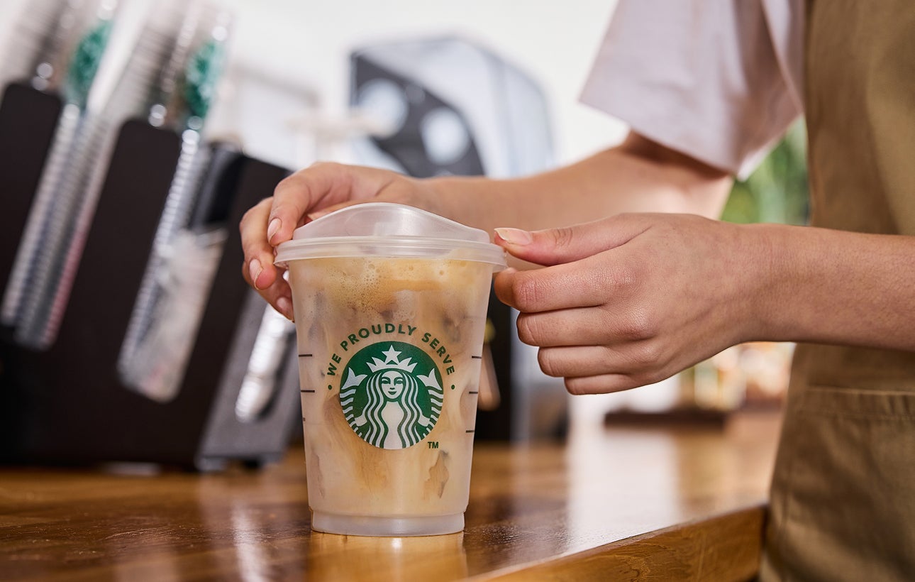 Un barista servant un verre We Proudly Serve Starbucks rempli de café glacé, dévoilant des couches de café et de crème