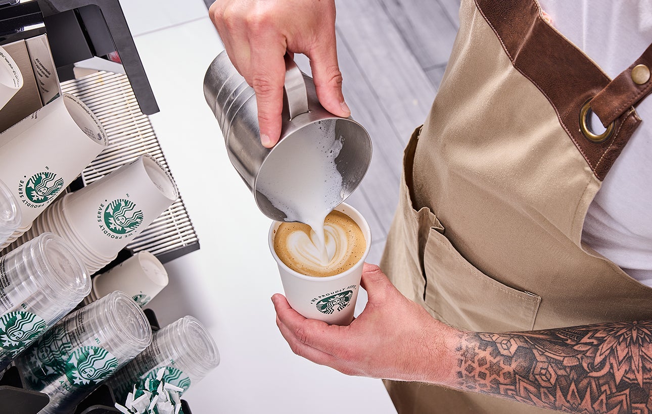 Un barista servant un verre We Proudly Serve Starbucks rempli de café glacé, dévoilant des couches de café et de crème