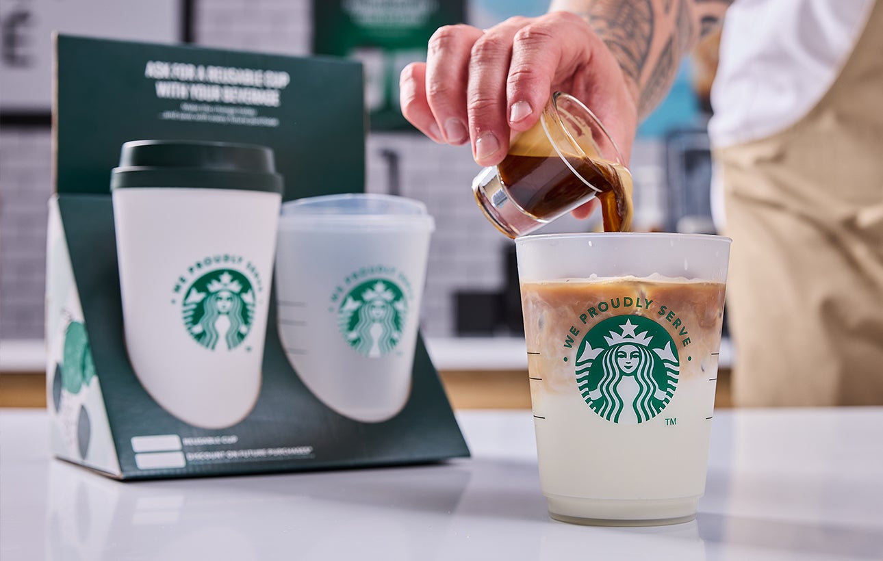 Un barista servant un verre We Proudly Serve Starbucks rempli de café glacé, dévoilant des couches de café et de crème