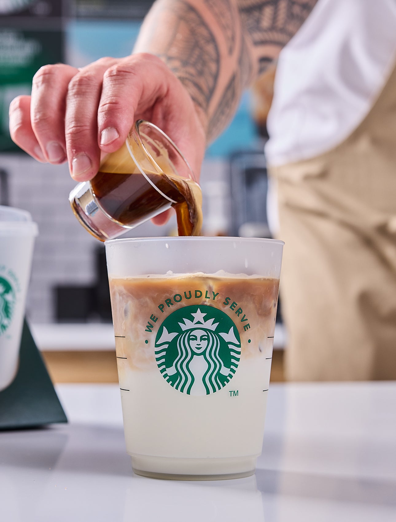 Un barista servant un verre We Proudly Serve Starbucks rempli de café glacé, dévoilant des couches de café et de crème
