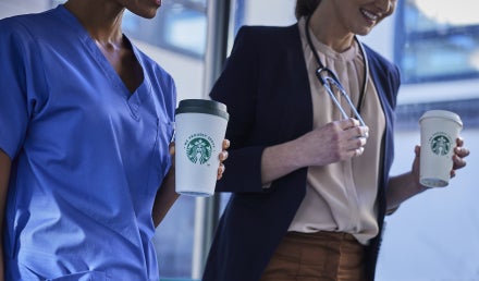 Deux professionnels de la santé marchent ensemble dans un établissement médical, souriants et tenant des boissons We Proudly 