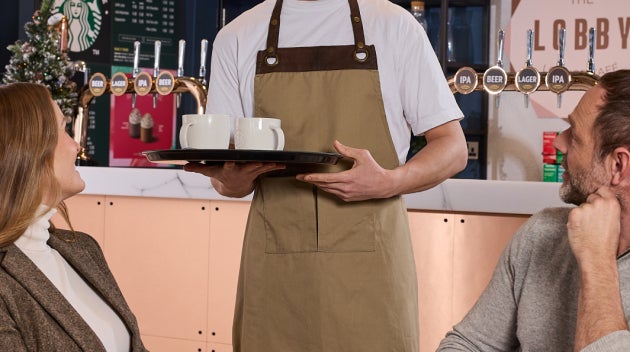 Un barista sert deux boissons Starbucks à des clients dans le bar du hall d’un hôtel, avec un menu We Proudly Serve Starbucks