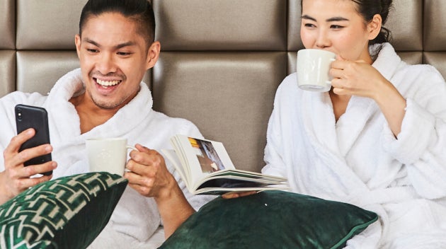 Un couple se détend sur un lit d’hôtel en peignoirs blancs, savourant un café Starbucks. 