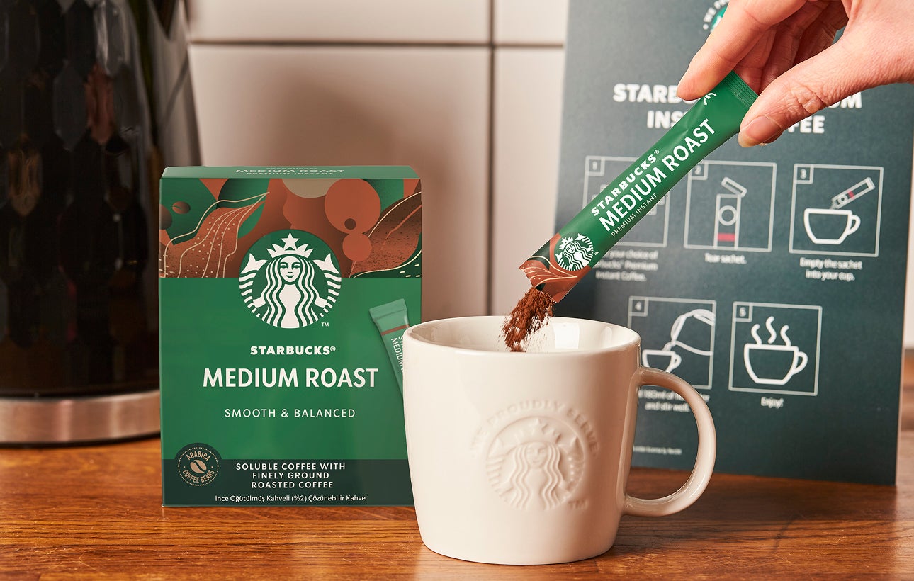 A Starbucks Premium Instant Medium Roast coffee sachet being poured into a ceramic We Proudly Serve Starbucks mug