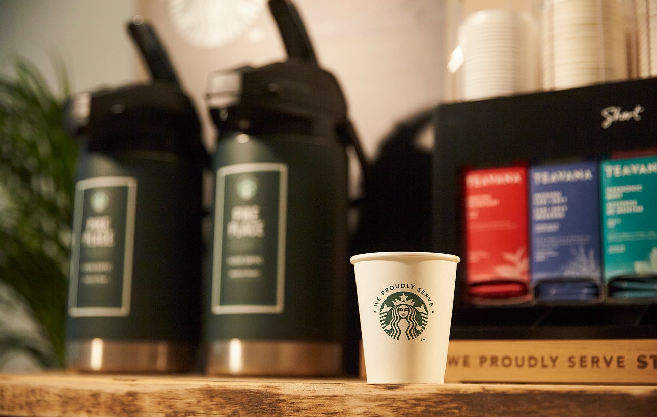 A We Proudly Serve Starbucks paper cup sits on a wooden counter in front of large brewed coffee dispensers