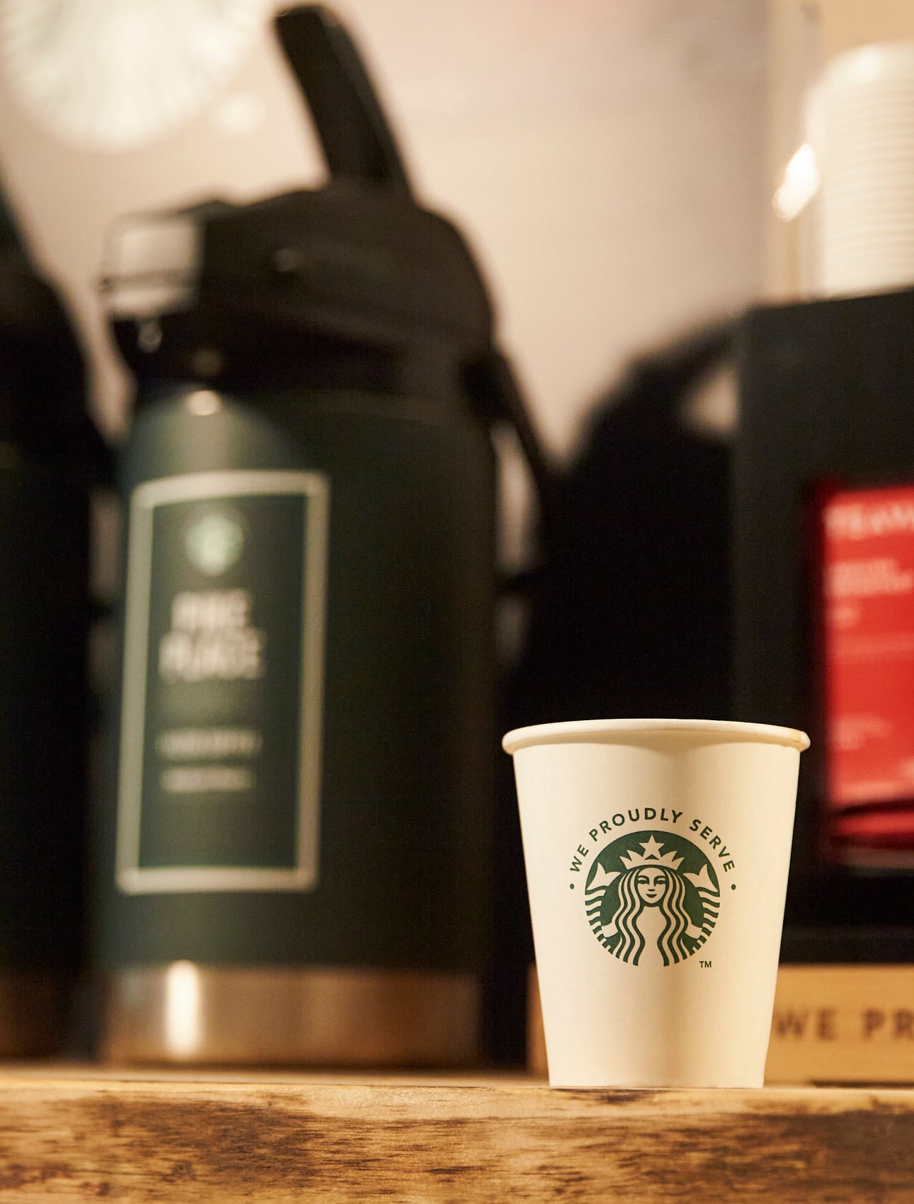 A We Proudly Serve Starbucks paper cup sits on a wooden counter in front of large brewed coffee dispensers