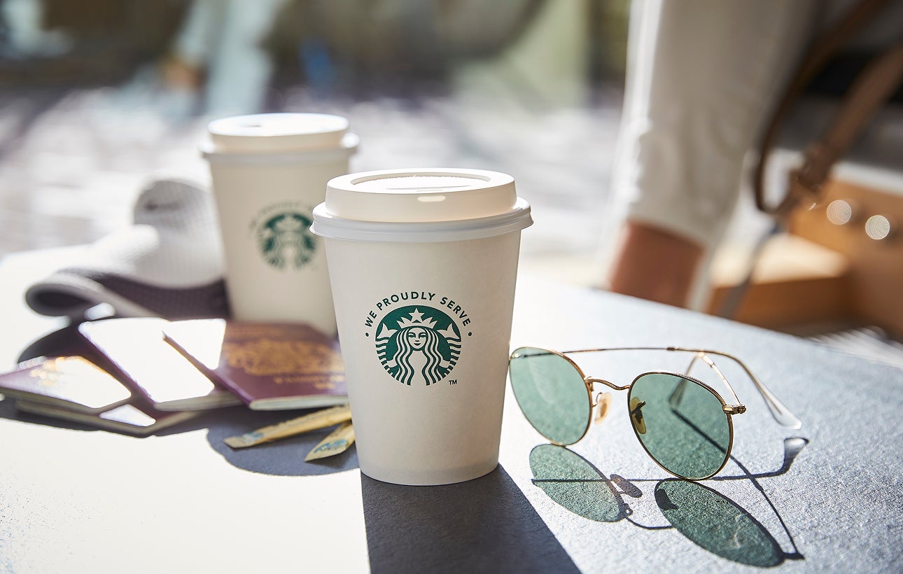 Two We Proudly Serve Starbucks takeaway cups with lids placed on an outdoor table beside a pair of sunglasses