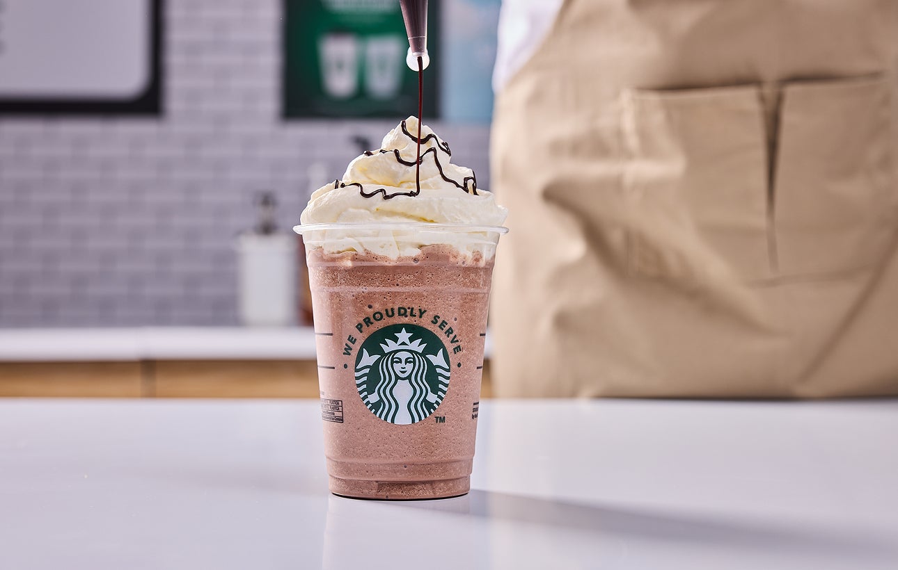 A We Proudly Serve Starbucks beverage topped with whipped cream, with a barista adding chocolate drizzle on top