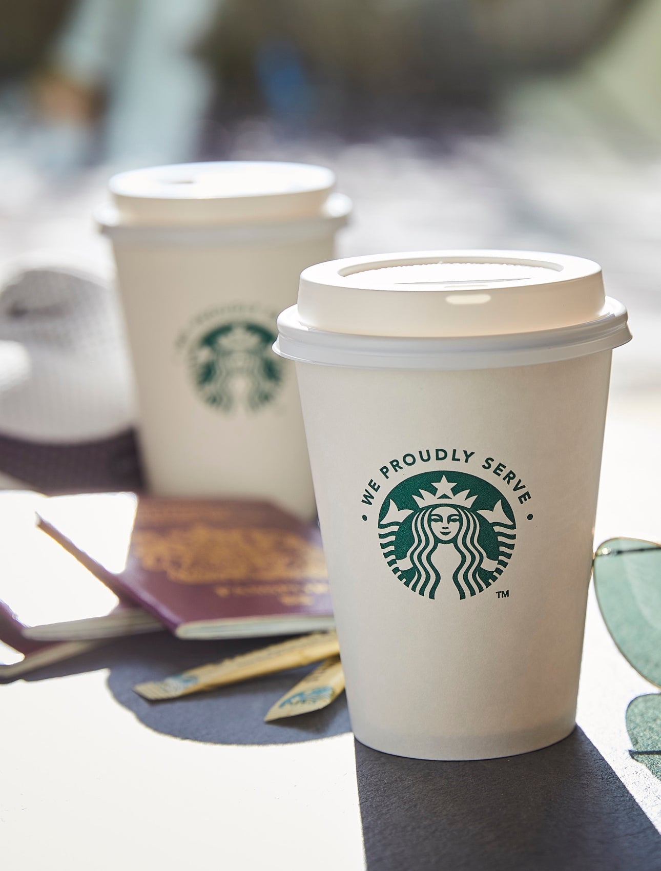 Two We Proudly Serve Starbucks takeaway cups with lids placed on an outdoor table beside a pair of sunglasses