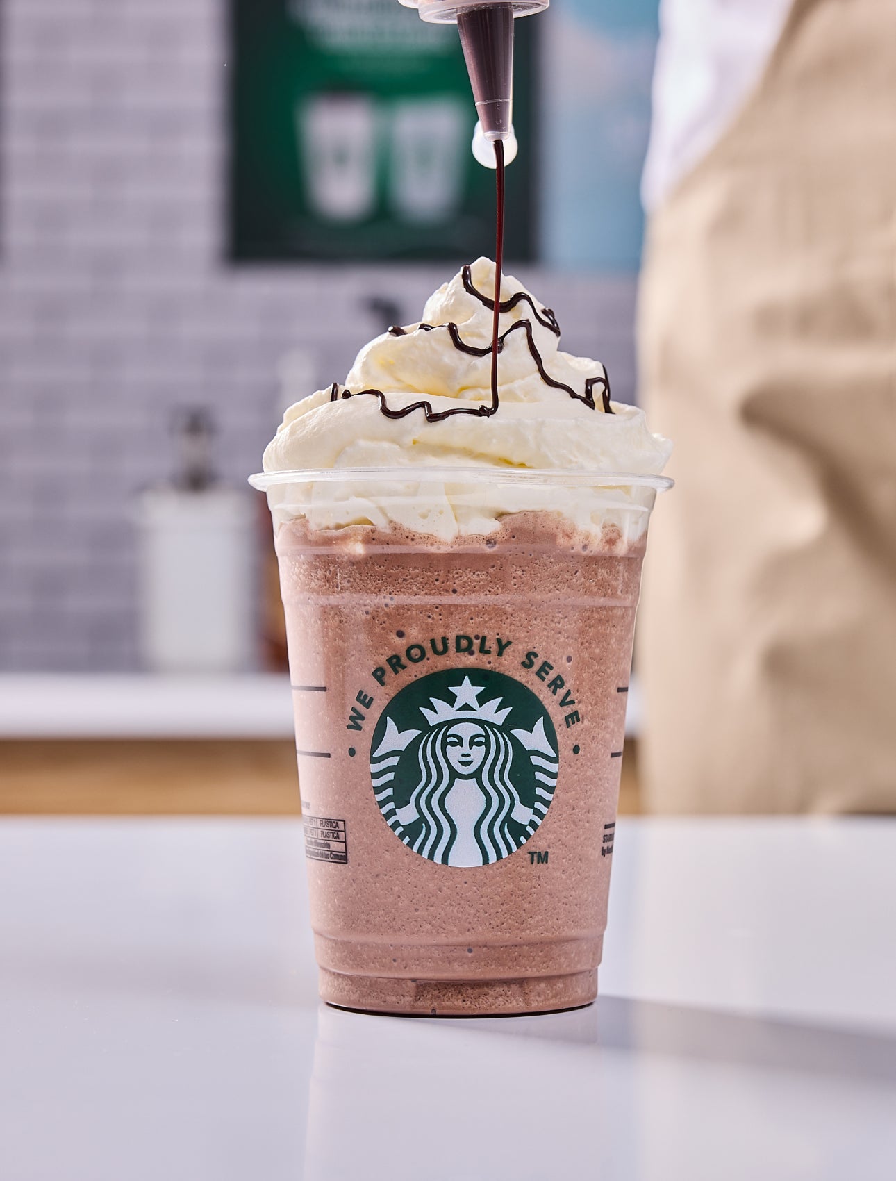 A We Proudly Serve Starbucks beverage topped with whipped cream, with a barista adding chocolate drizzle on top