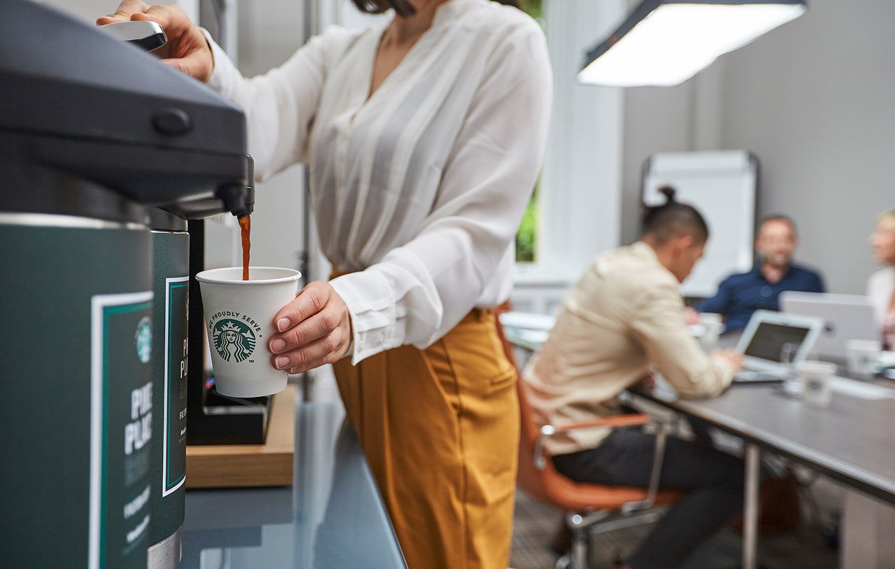 A person fills a We Proudly Serve Starbucks paper cup with brewed coffee from a commercial coffee dispenser