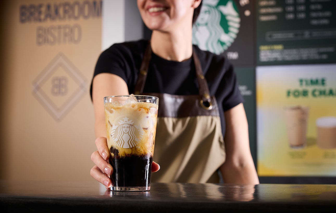 A barista serving a We Proudly Serve Starbucks glass filled with iced coffee, showcasing layers of coffee and cream