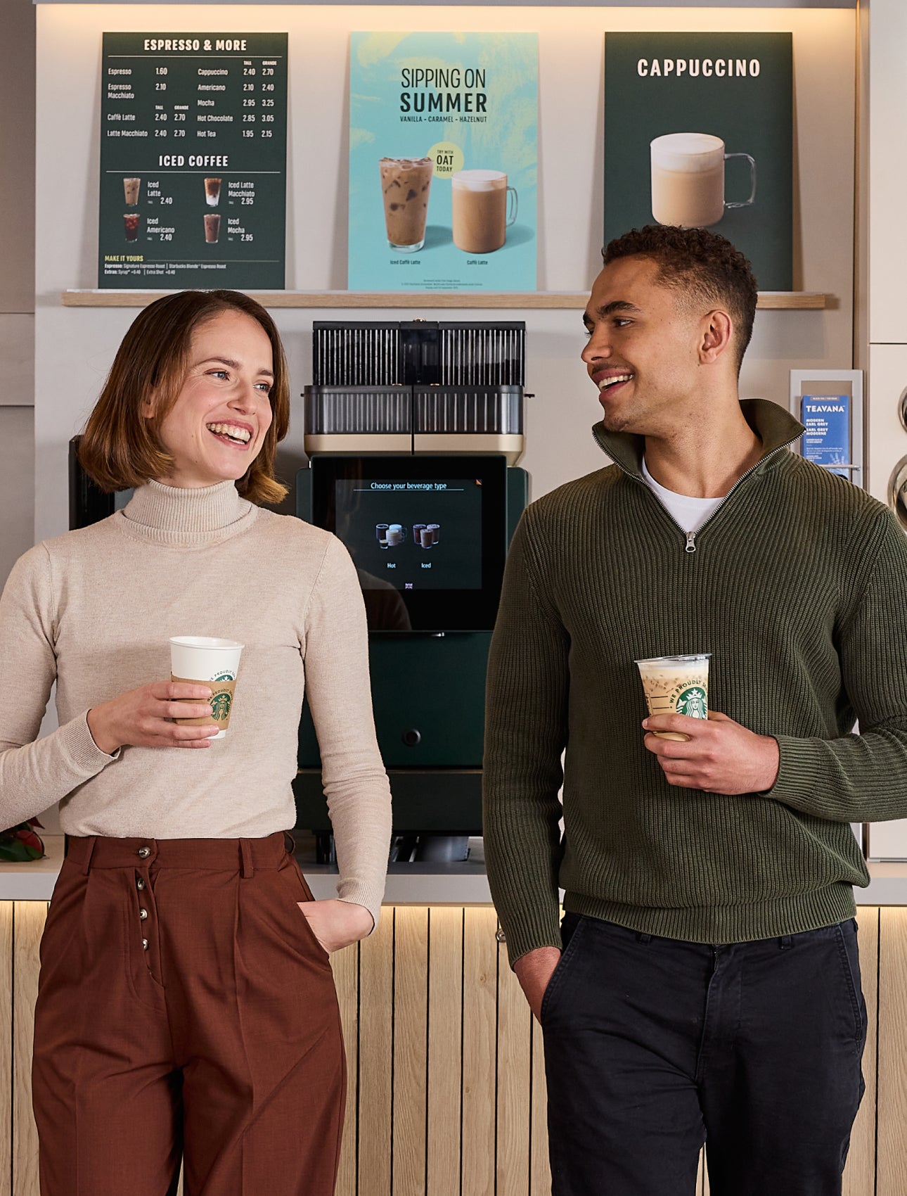 Two people standing and chatting while holding We Proudly Serve Starbucks coffee cups in front of a self-serve coffee station