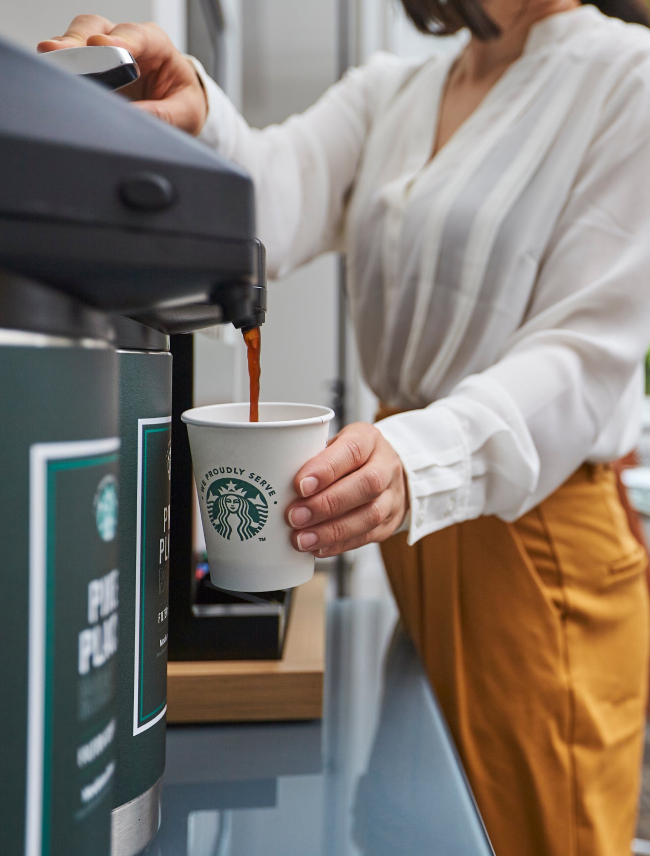 A person fills a We Proudly Serve Starbucks paper cup with brewed coffee from a commercial coffee dispenser