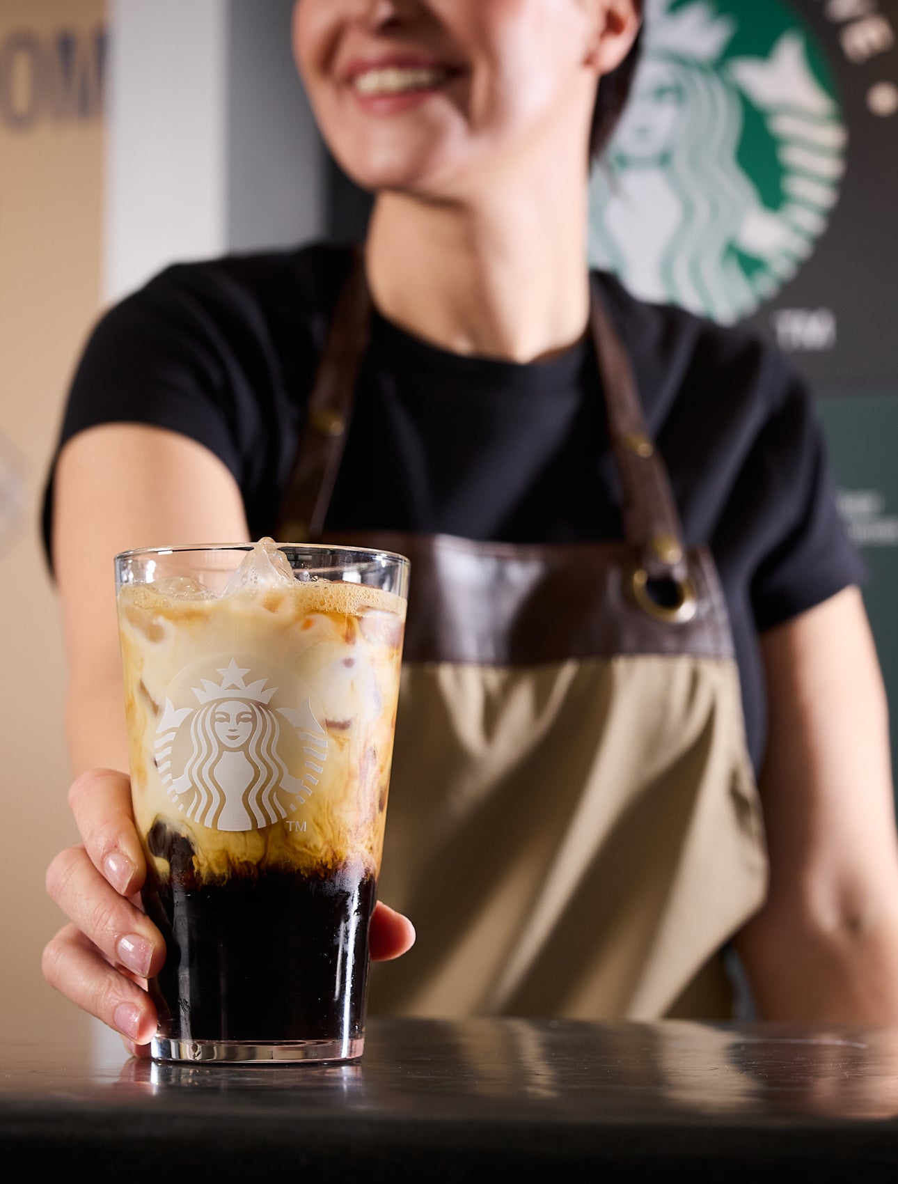 A barista serving a We Proudly Serve Starbucks glass filled with iced coffee, showcasing layers of coffee and cream