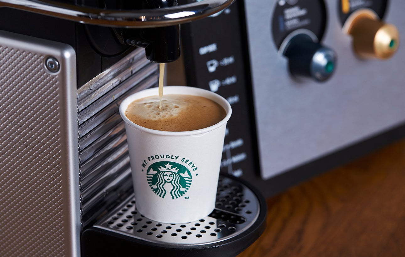 A We Proudly Serve Starbucks paper cup being filled with freshly brewed coffee from an espresso machine