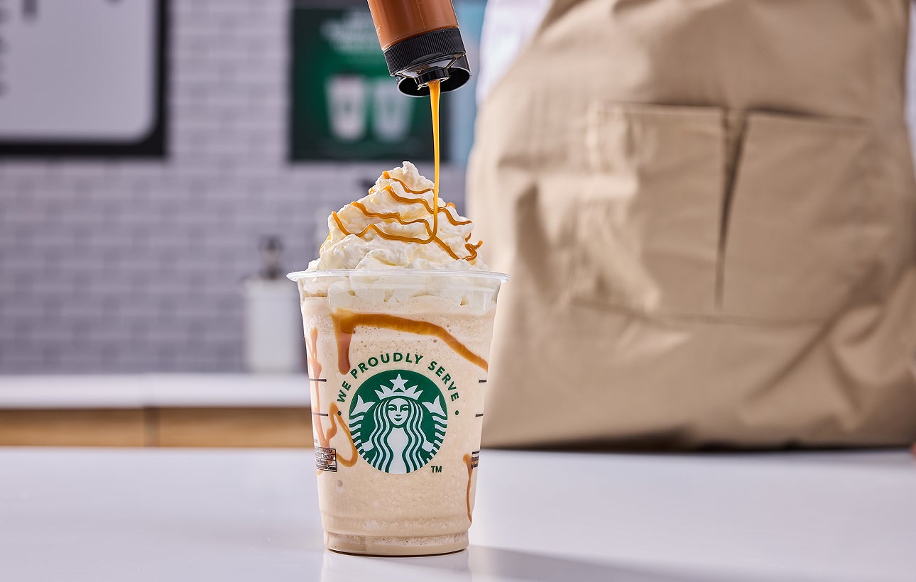 A Starbucks blended drink topped with whipped cream, with a barista adding caramel drizzle