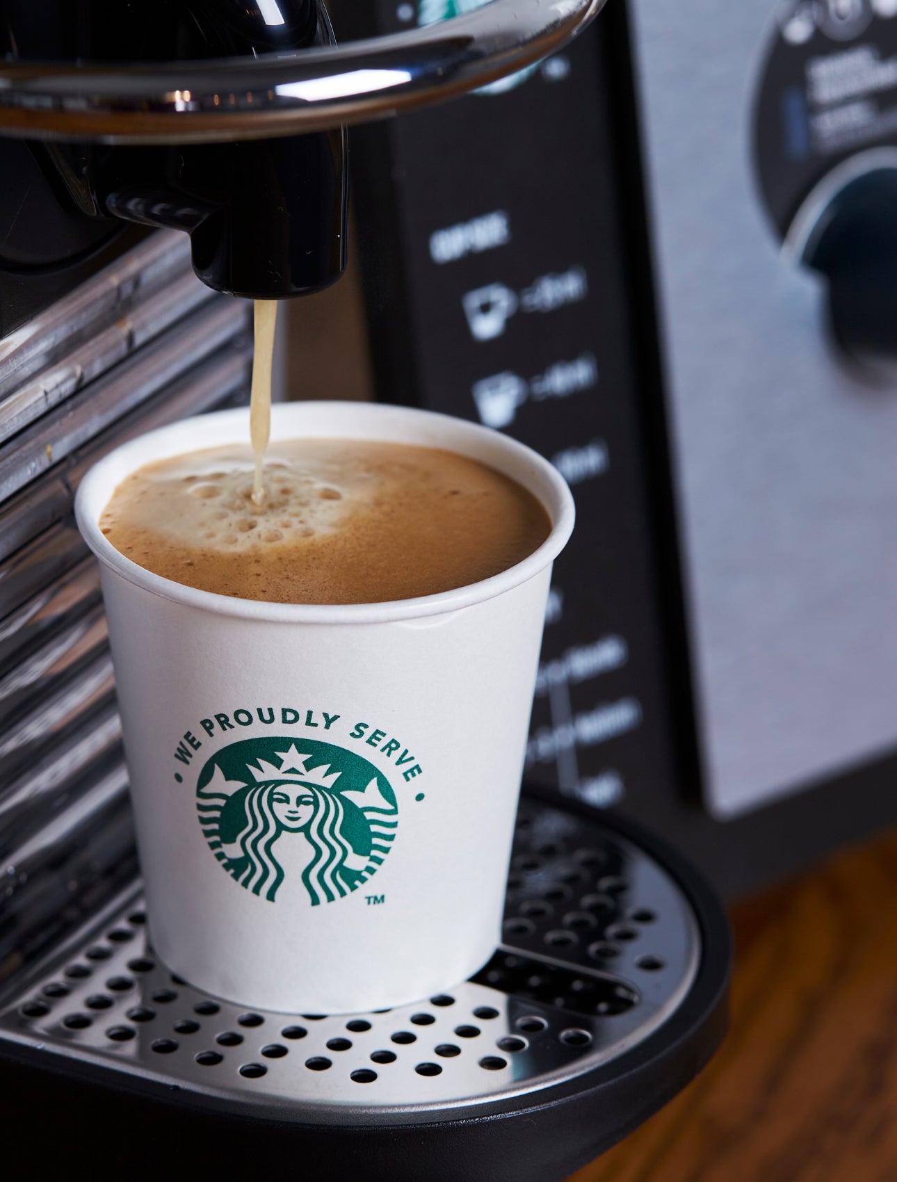 A We Proudly Serve Starbucks paper cup being filled with freshly brewed coffee from an espresso machine