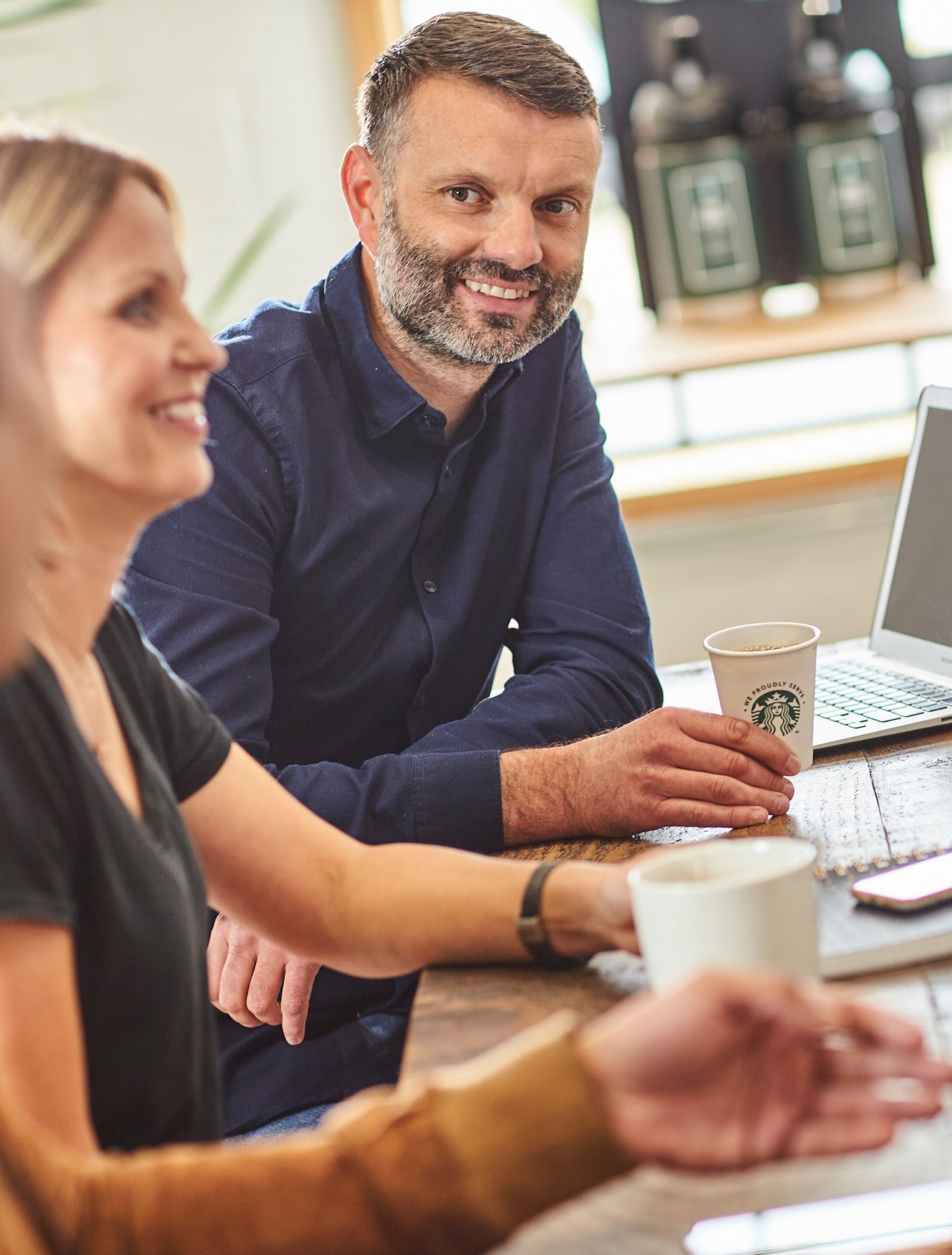 A group of people sitting at a table, smiling and talking, with a cup of brewed coffee beside a laptop