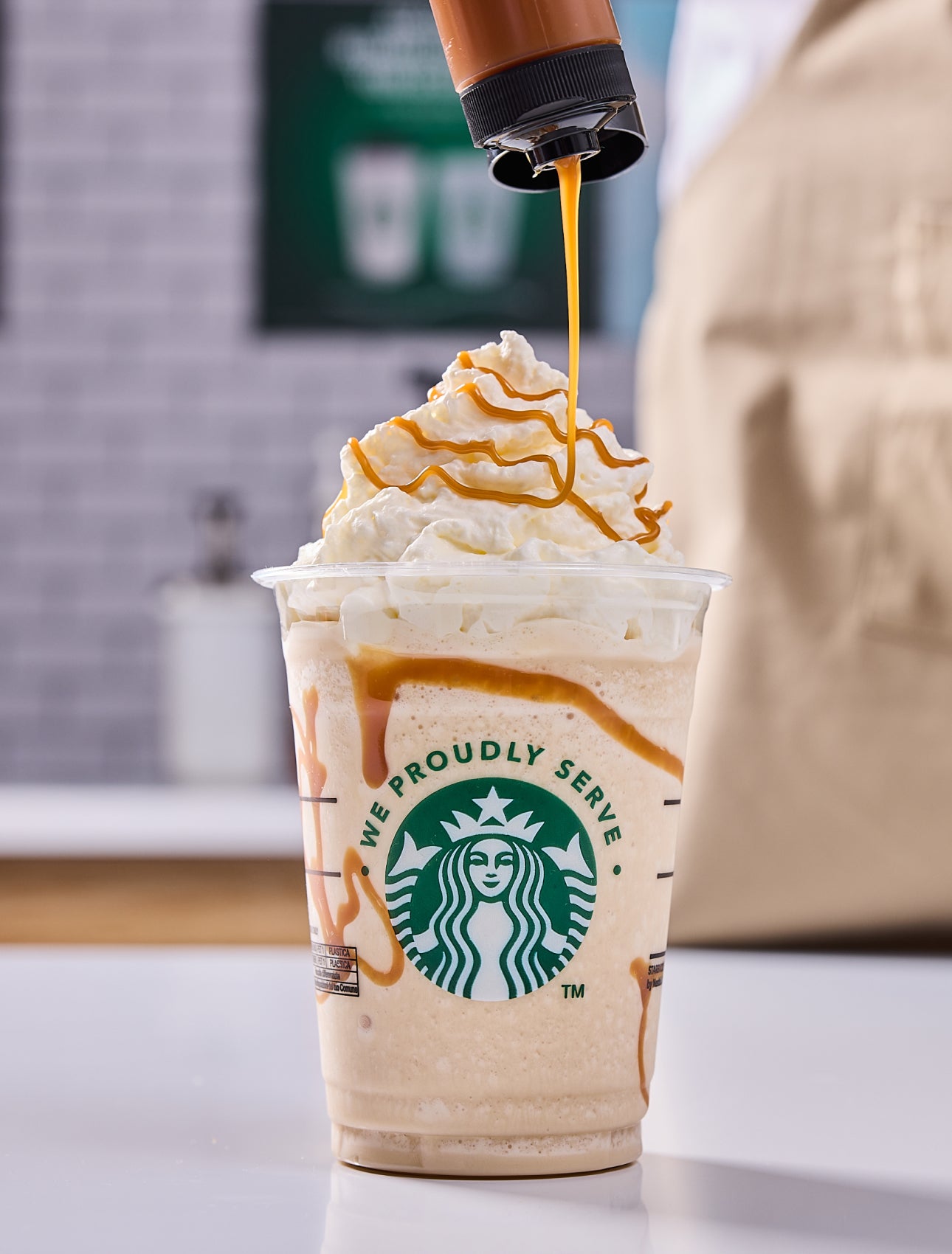 A Starbucks blended drink topped with whipped cream, with a barista adding caramel drizzle