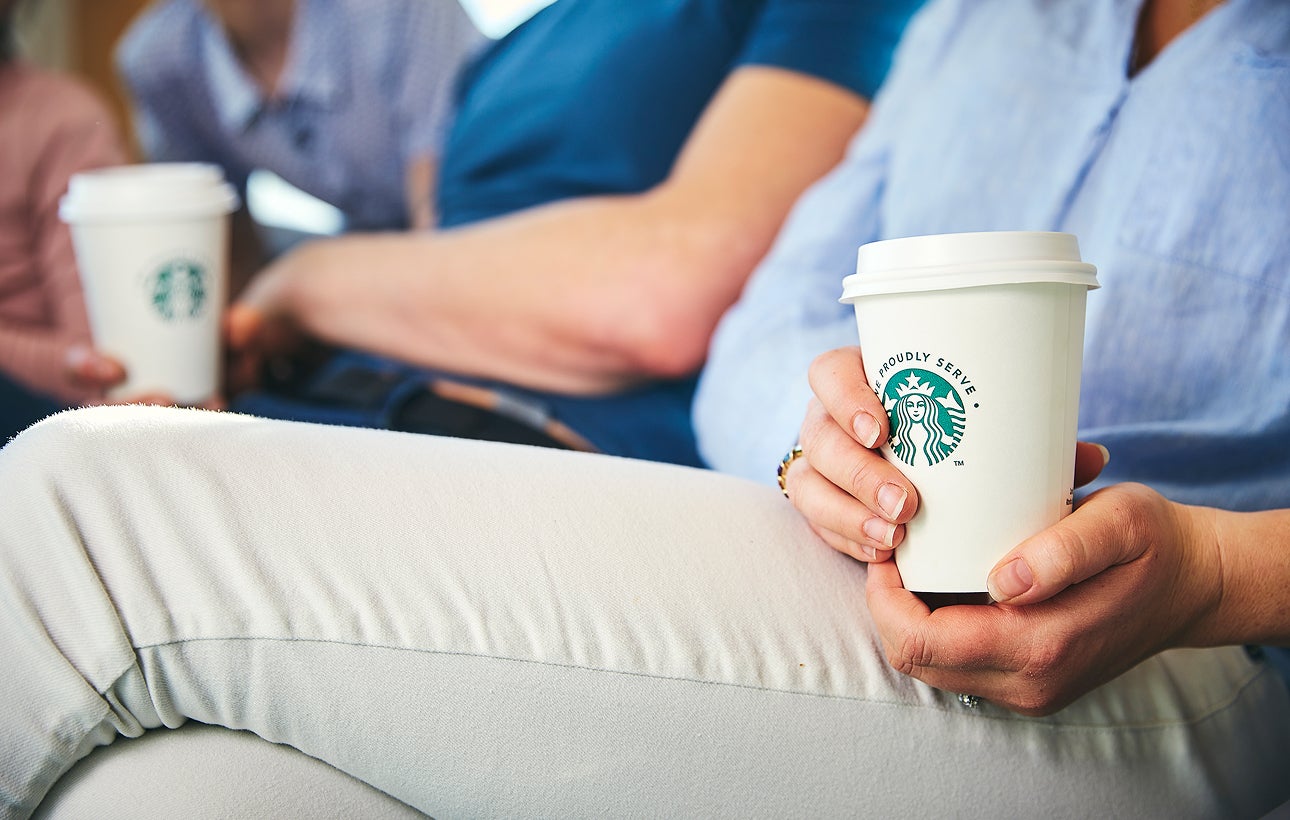 A person sitting down, holding a We Proudly Serve Starbucks takeaway cup with both hands