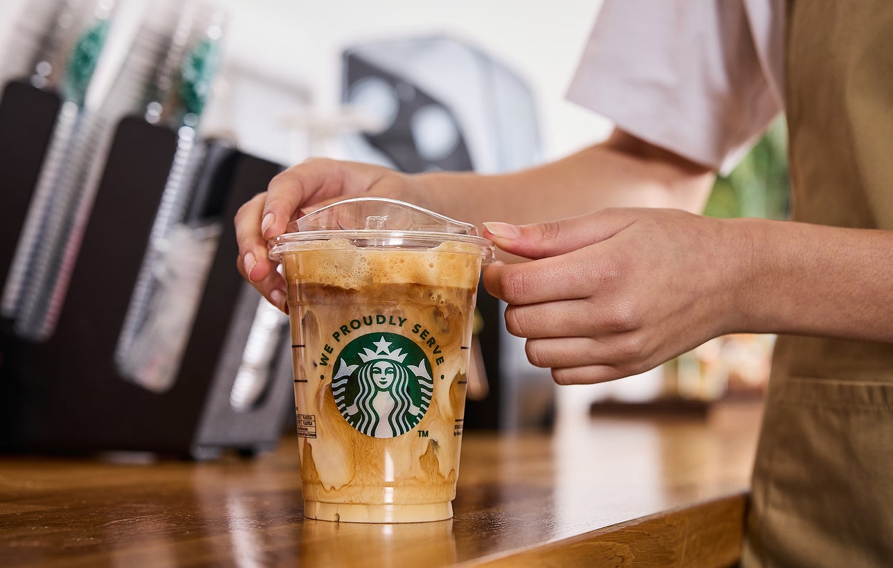 A barista places a lid on a We Proudly Serve Starbucks cup of iced coffee, featuring swirls of cream and coffee