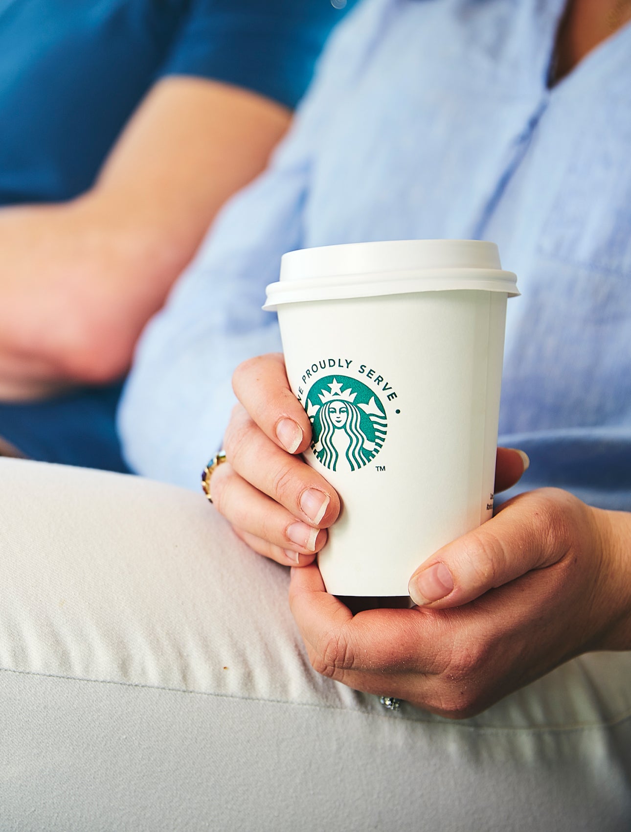 A person sitting down, holding a We Proudly Serve Starbucks takeaway cup with both hands