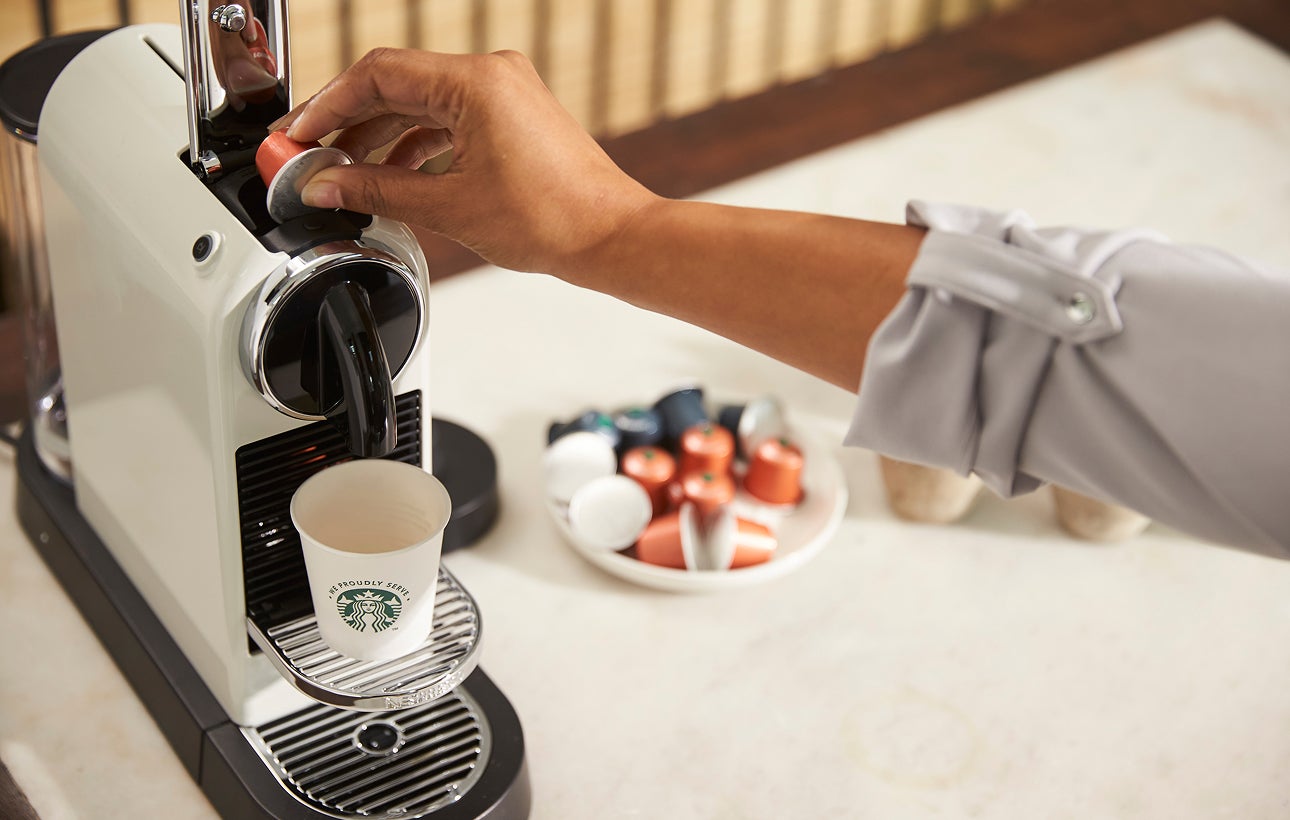 A person inserts a coffee capsule into a single-serve coffee machine, with a We Proudly Serve Starbucks cup on the drip tray