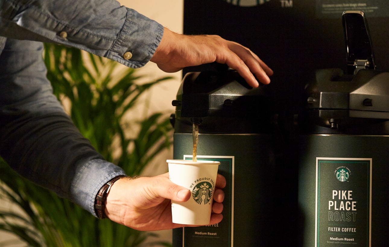 A person dispensing Starbucks Pike Place® Roast coffee from a large coffee dispenser into a We Proudly Serve Starbucks cup