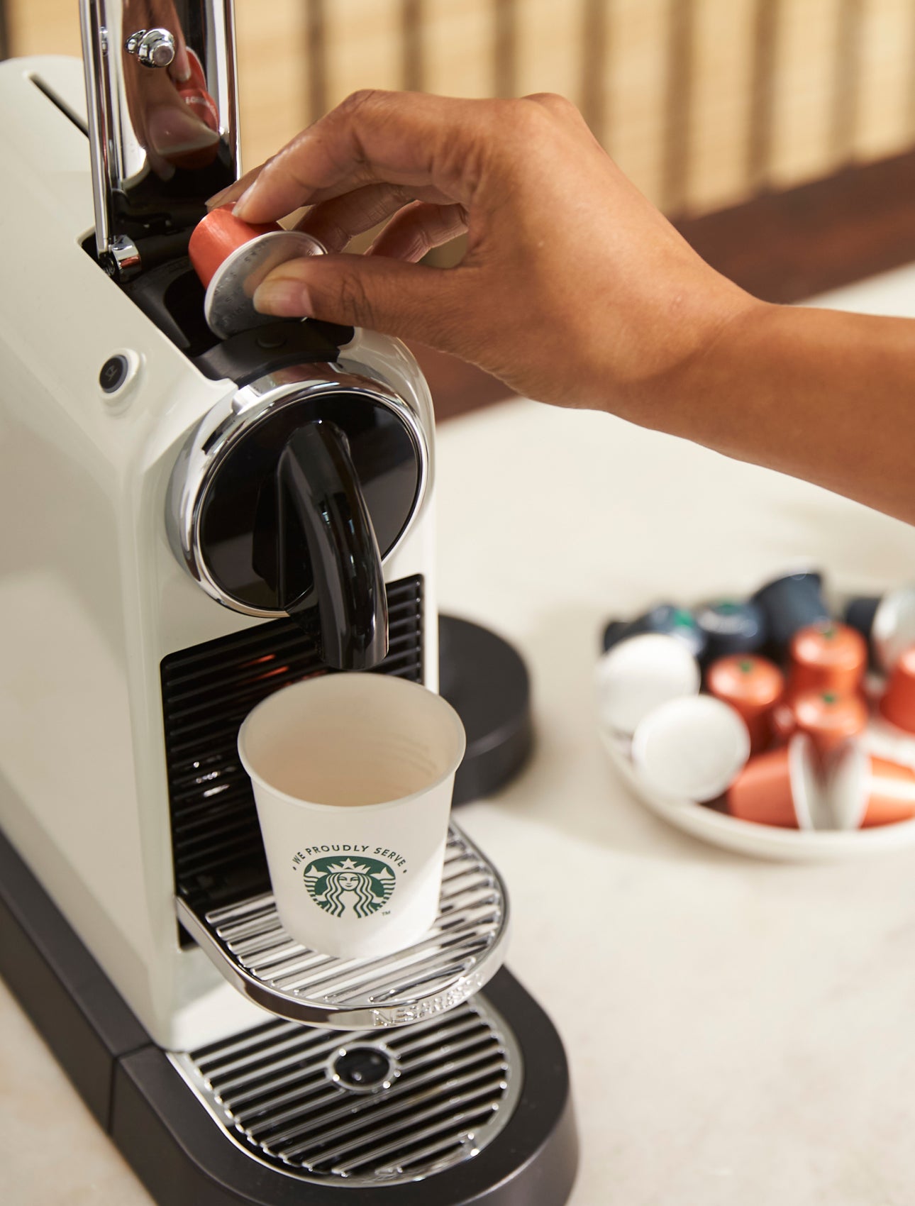 A person inserts a coffee capsule into a single-serve coffee machine, with a We Proudly Serve Starbucks cup on the drip tray