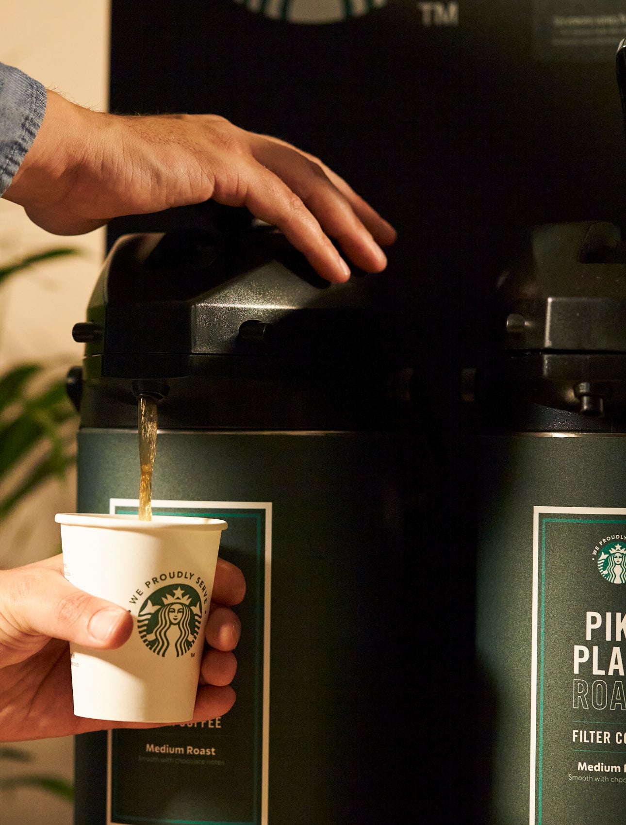 A person dispensing Starbucks Pike Place® Roast coffee from a large coffee dispenser into a We Proudly Serve Starbucks cup