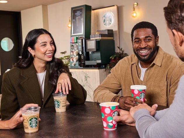 Four people at a café table, smiling and holding Starbucks drinks in festive and standard We Proudly Serve Starbucks cups