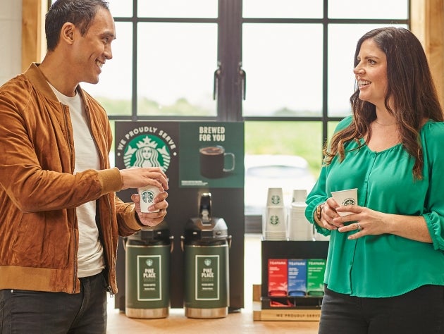 Two people standing and chatting while holding We Proudly Serve Starbucks cups in front of brewed coffee dispensers