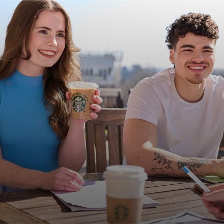 A young man and woman sitting outside on a sunny day, smiling and talking while holding We Proudly Serve Starbucks beverages