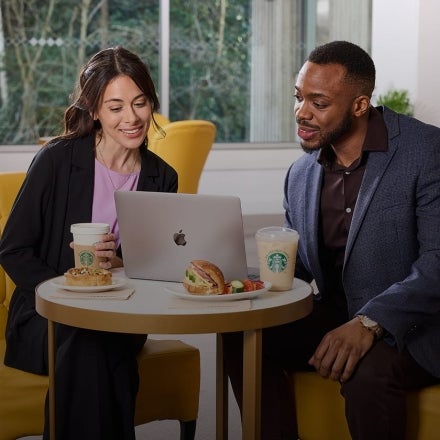 Two people sitting in front of a laptop with We Proudly Serve Starbucks coffee and sandwiches on the table