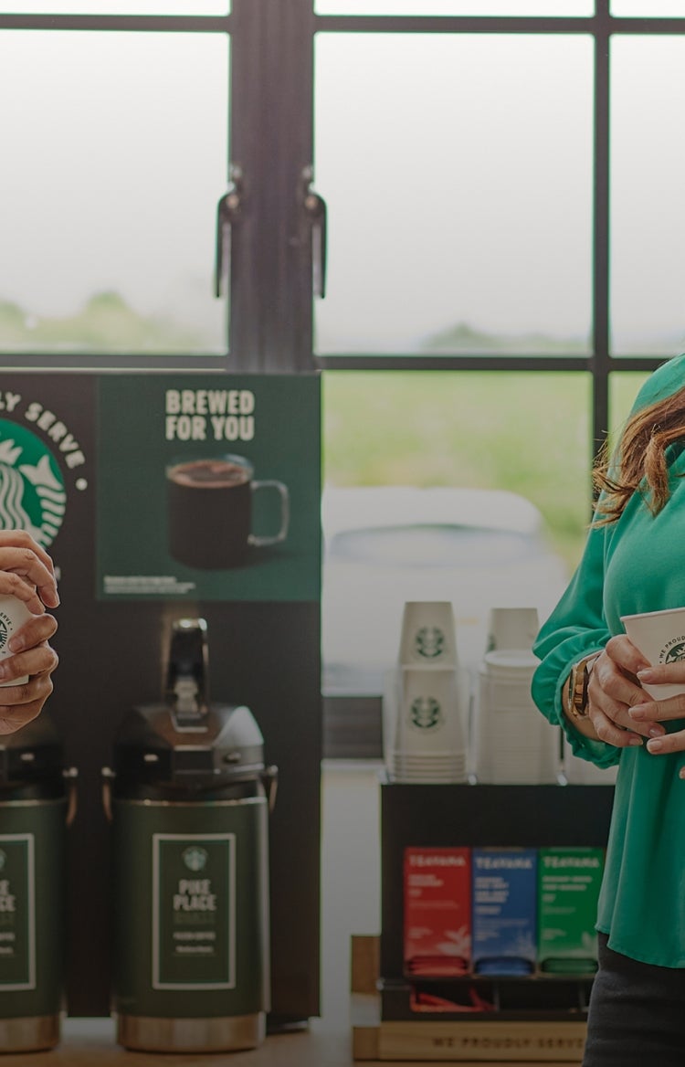 Two people standing and chatting while holding We Proudly Serve Starbucks cups in front of brewed coffee dispensers