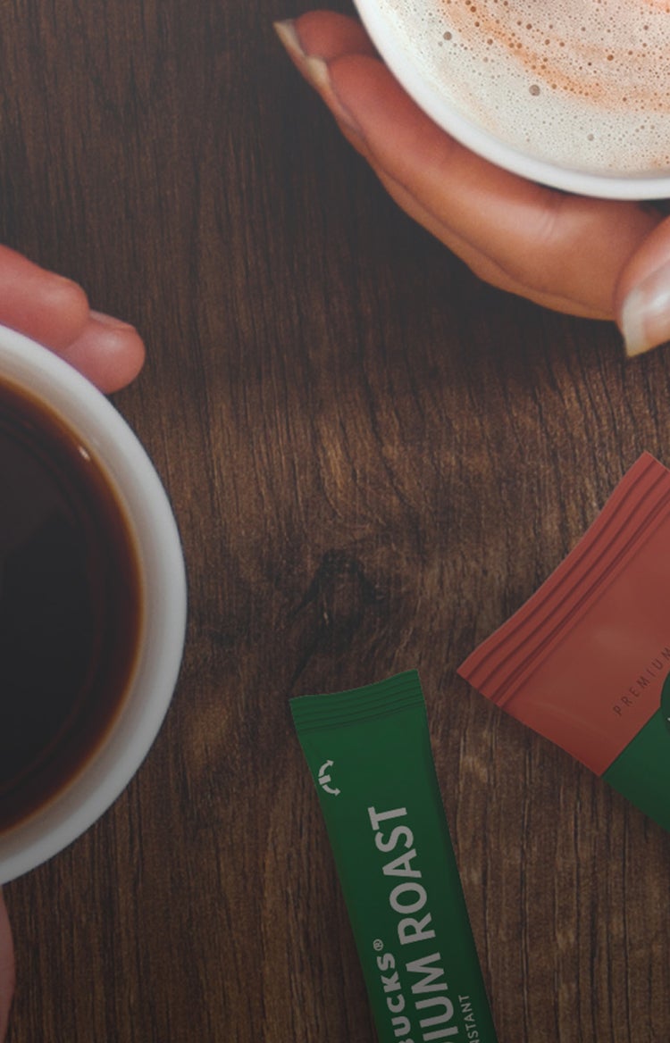 Two people holding coffee cups over a wooden table, with Starbucks cappuccino and Colombia roast sachets nearby.