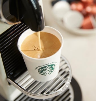A We Proudly Serve Starbucks cup being filled with freshly brewed coffee from a single-serve espresso machine