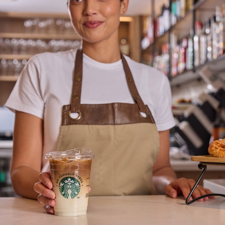 A barista serving a transparent We Proudly Serve Starbucks cup of iced coffee