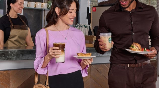 A man and woman standing at a workplace café counter, smiling as they pick up Starbucks iced coffee and food