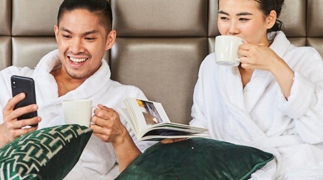 A couple relaxing on a hotel bed in white robes, enjoying Starbucks coffee. 