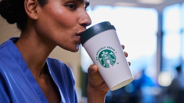 A healthcare worker in blue scrubs drinking a We Proudly Serve Starbucks coffee while sitting in a hospital staff restaurant