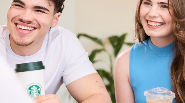 A young man and woman laughing together while sitting at a table, each holding a We Proudly Serve Starbucks drink