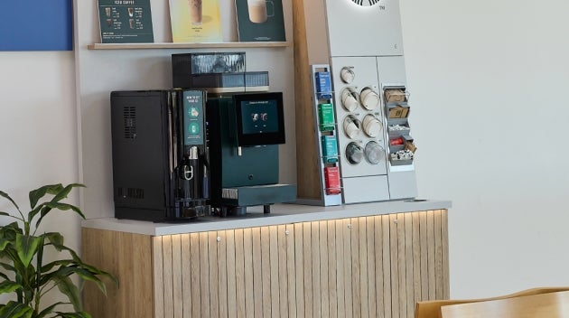 A compact We Proudly Serve Starbucks coffee station in a hospital outpatient area, featuring a self-serve coffee machine