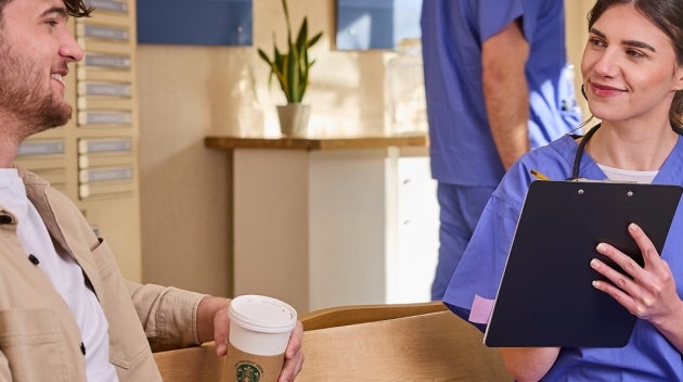 A woman in blue scrubs talking to a patient holding a We Proudly Serve Starbucks coffee cup in a clinic.